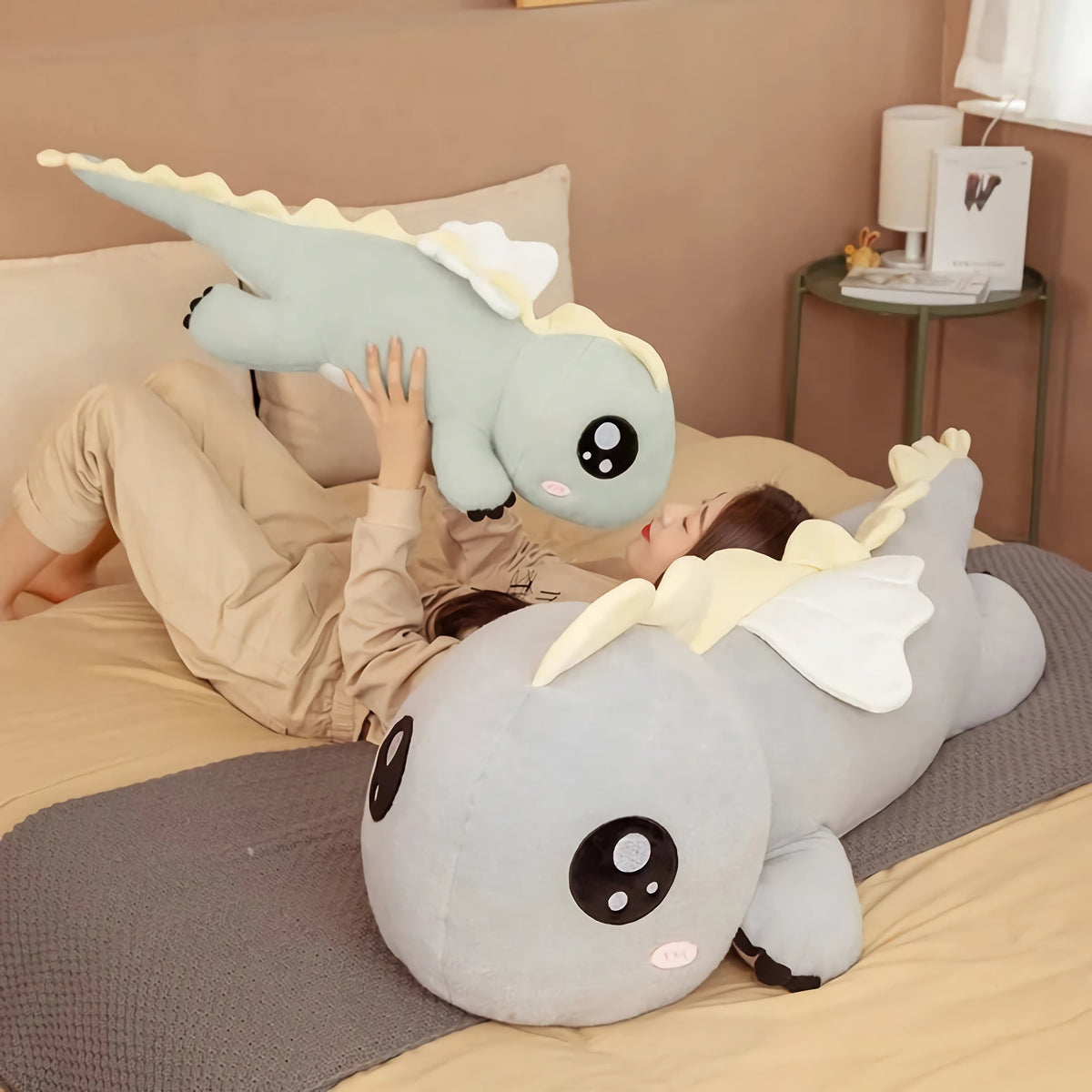 A child lies on a bed holding a small plush dinosaur while a larger plush dinosaur rests beside them
