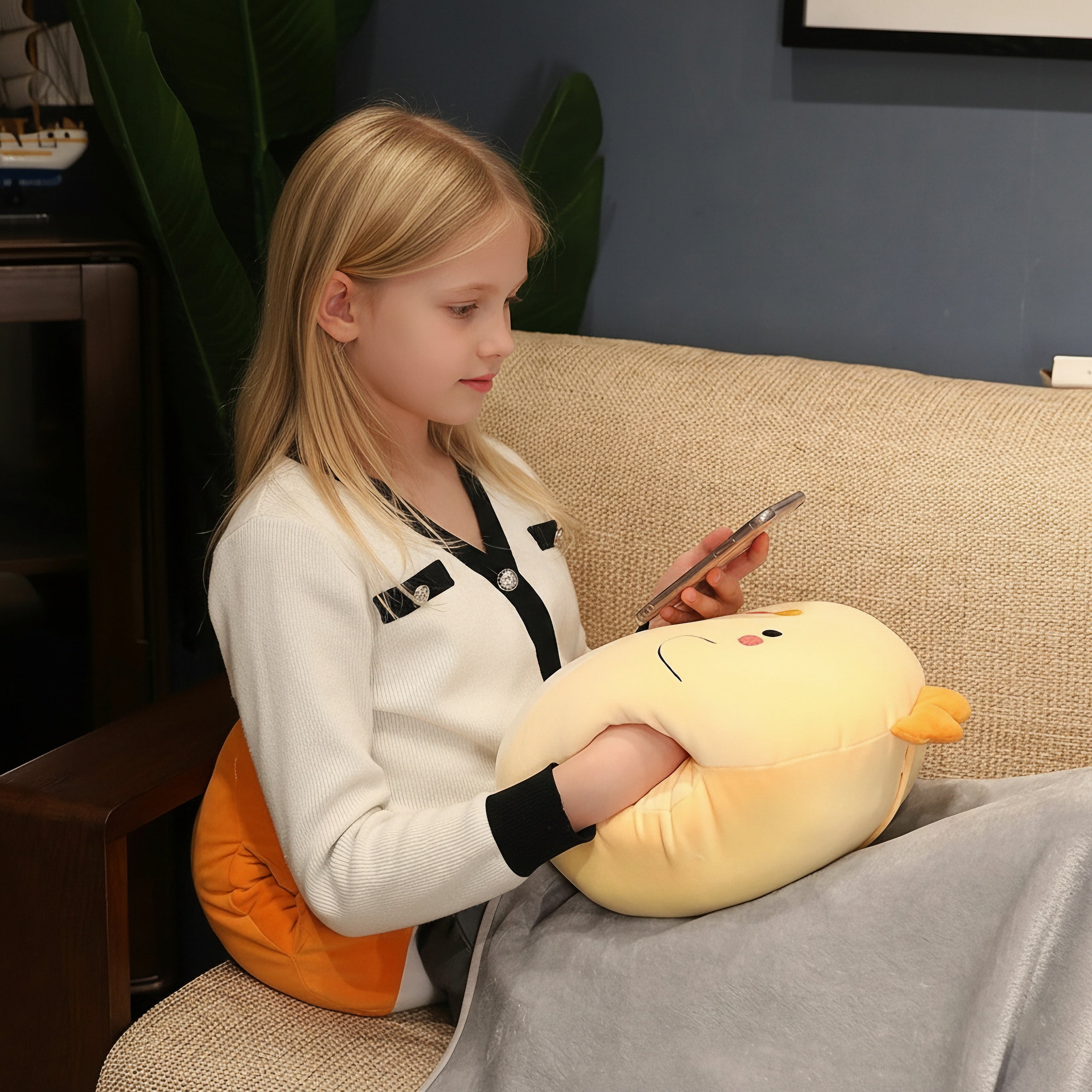 Child sitting on a couch with a folded travel blanket draped over her lap next to a plush pillow, showing how the 100cm and 150cm blanket-plush sizes compare in everyday use