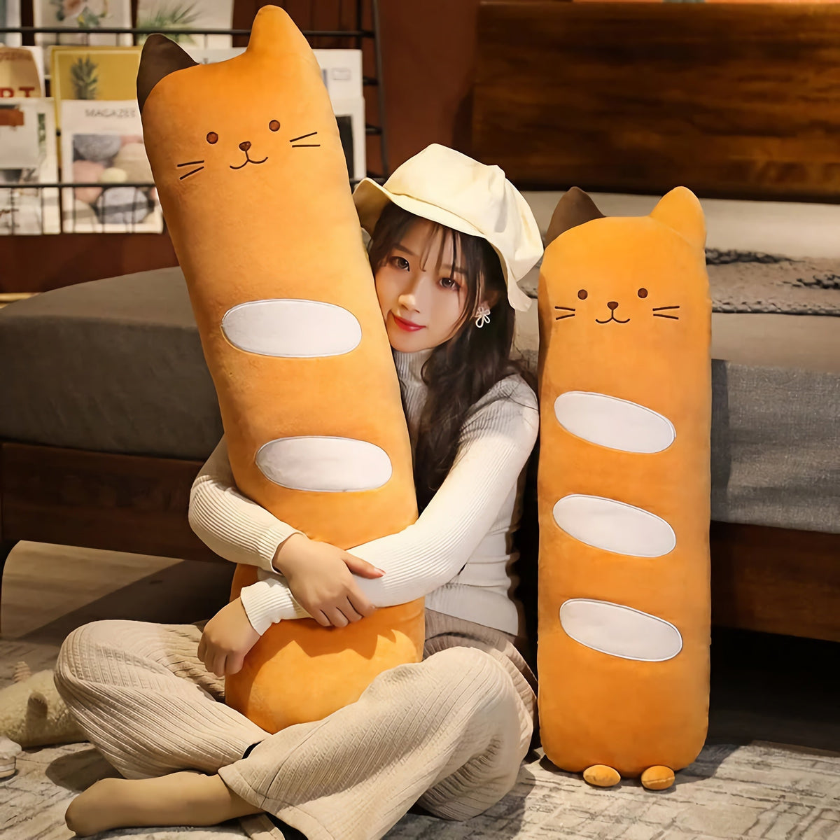 Person sitting on the floor hugging a large long cat bolster pillow plush, with a smaller one beside it