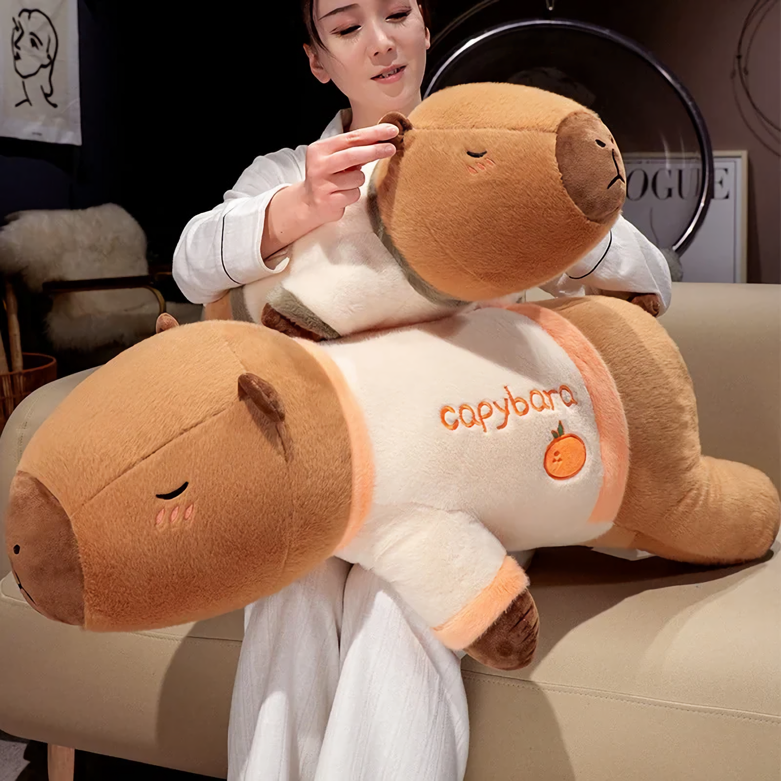 Woman sitting on a sofa lifting a lying capybara plush pillow in a cream shirt
