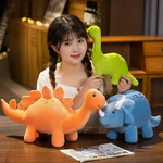 Woman holding a green Brontosaurus plush, with orange Stegosaurus and blue Triceratops plushes on the table.