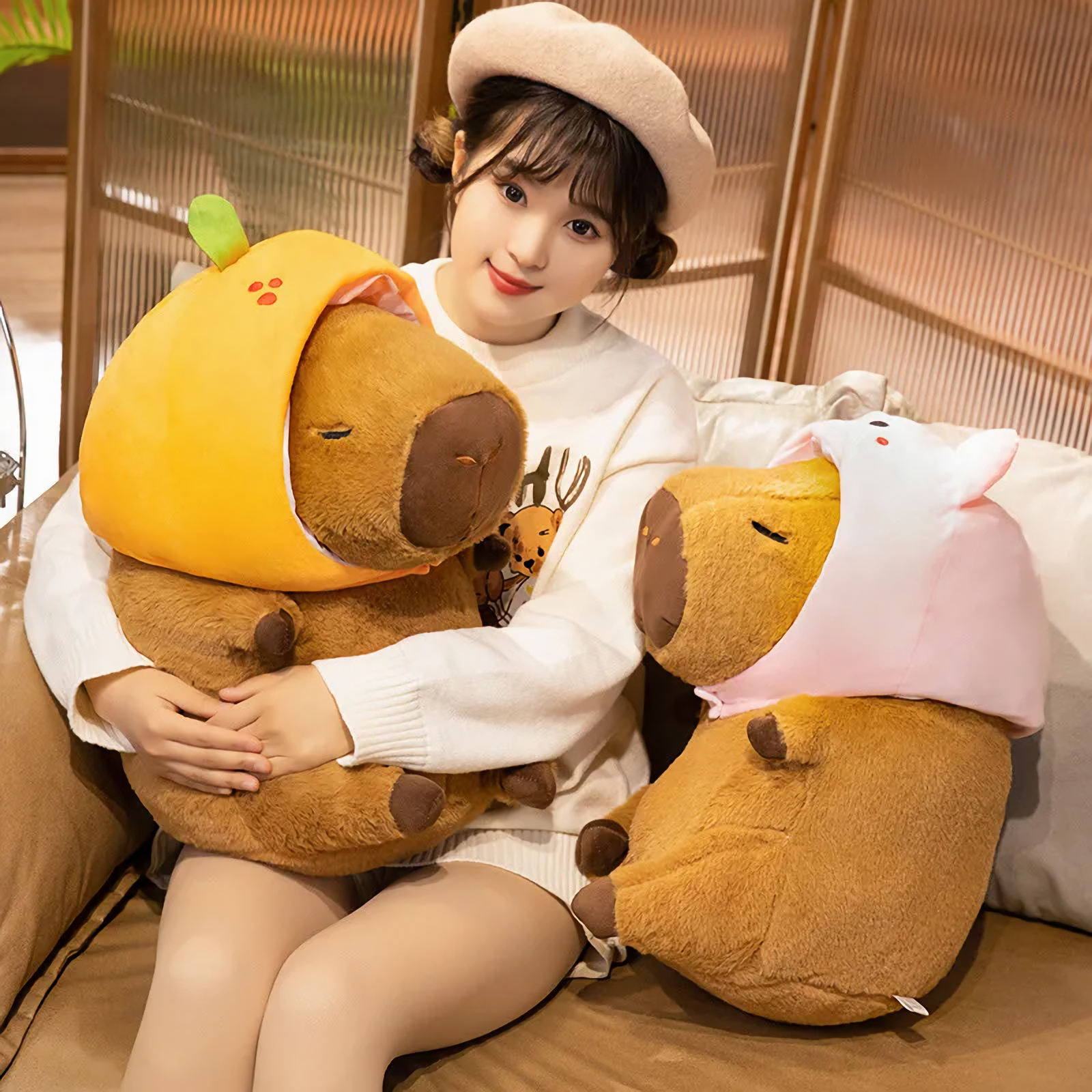 Woman hugging a capybara plush in an orange hood beside a pig hood capybara