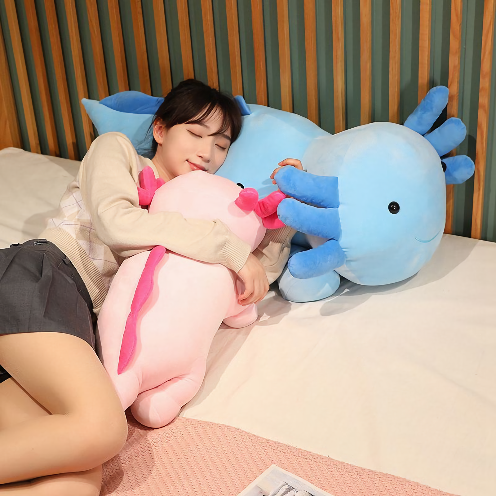 Girl lying on a bed hugging a pink axolotl plush pillow with a blue axolotl plush beside her