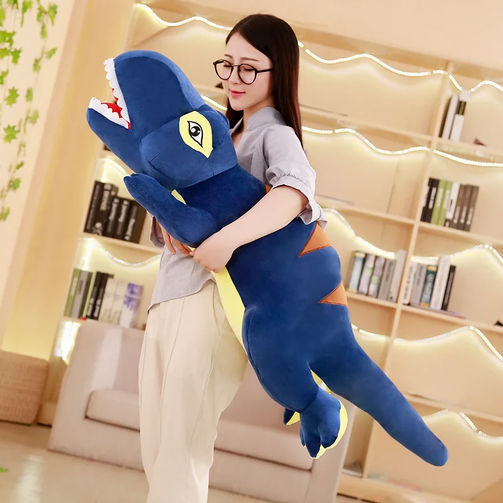 Woman with glasses hugging a long blue T-Rex plush pillow in a bright living room