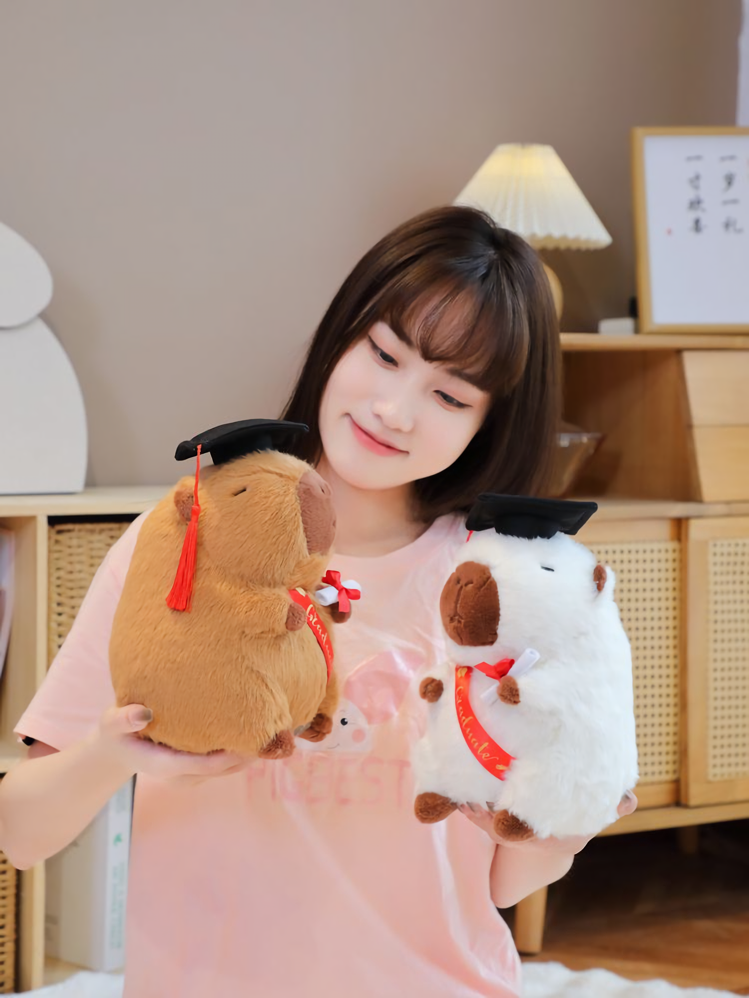 Woman holding a brown and a white graduation capybara plush with black caps
