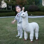 Young girl standing beside a white standing alpaca plush on a green lawn