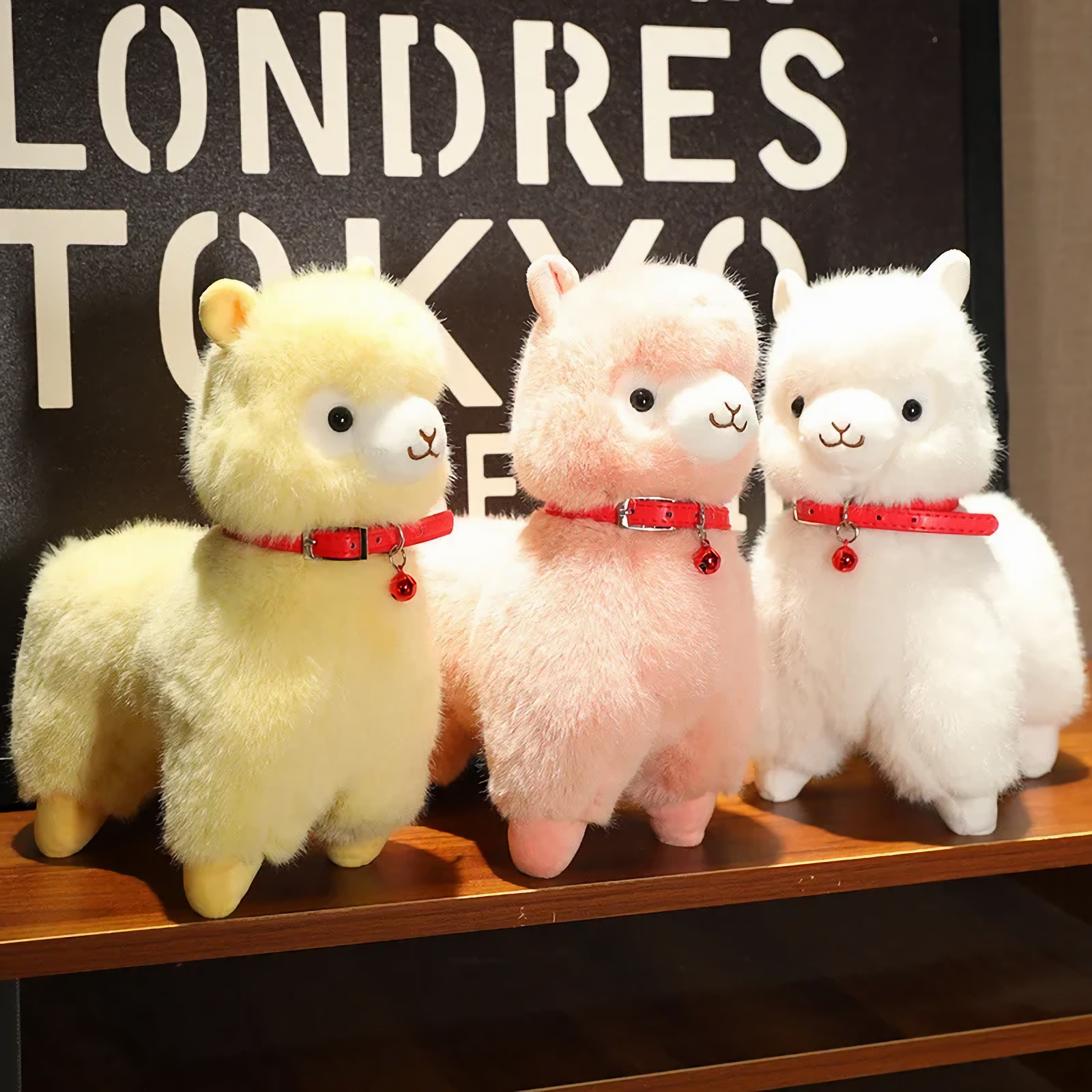 Yellow, pink and white alpaca plushies with red bell collars lined up on a wooden shelf