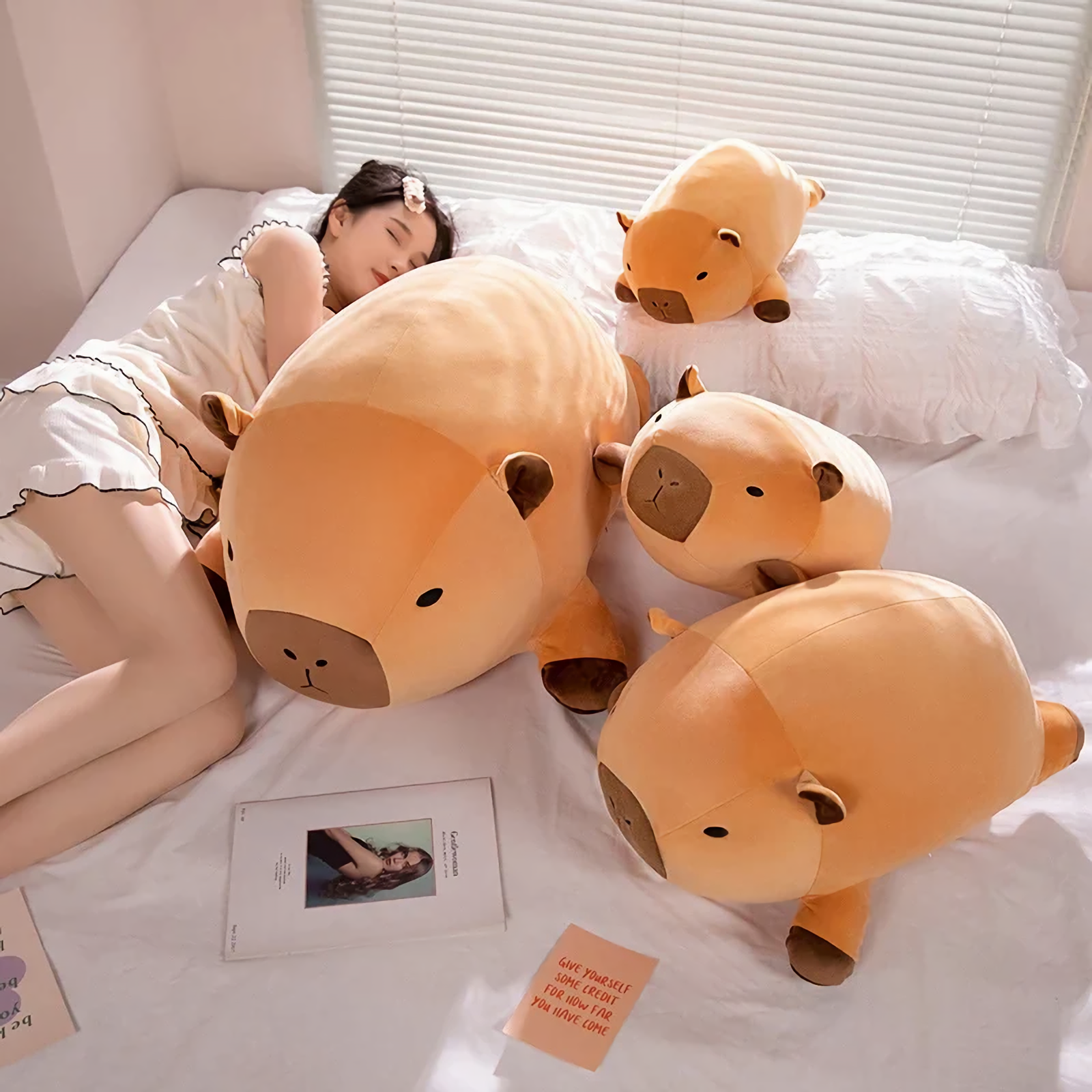 Woman lying on a bed beside three chubby capybara plush pillows of different sizes