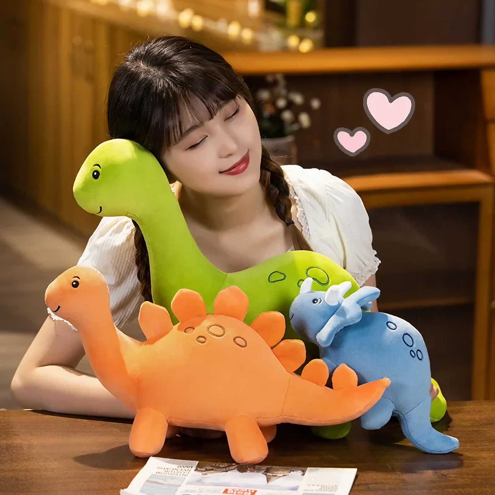 A woman hugs a green Brontosaurus plush, with orange Stegosaurus and blue Triceratops plushes on a table.