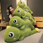 Woman on a bed with stacked green dinosaur plush pillows, brown and yellow spikes, comparing sizes.