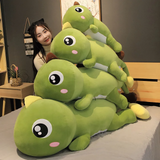 Woman on a bed with stacked green dinosaur plush pillows, brown and yellow spikes, comparing sizes.