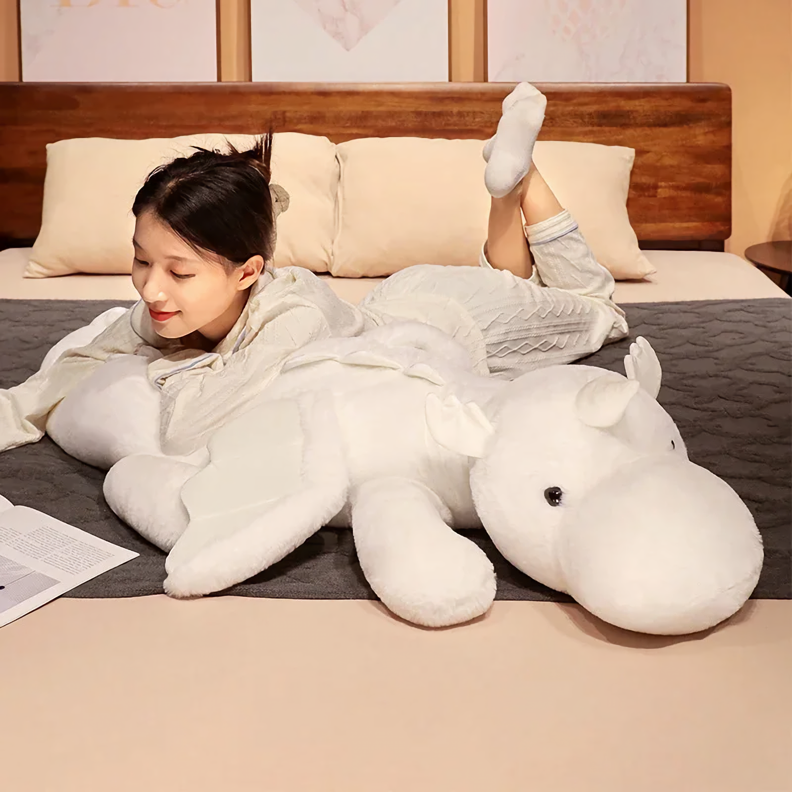Woman lying on bed hugging a white dragon plush body pillow with small wings and horns.