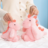 Hands holding two pink dinosaur plush toys on a white bed