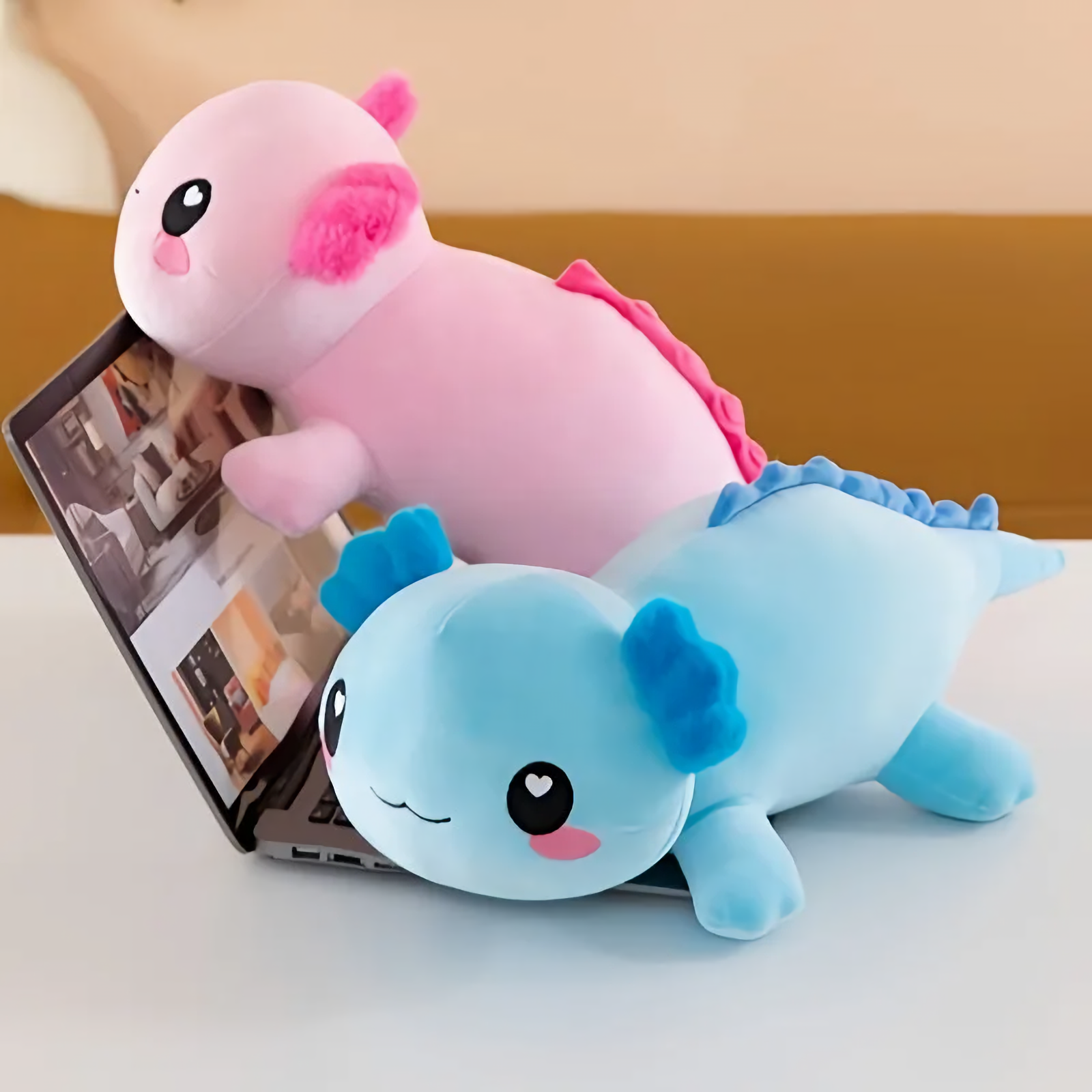 Pink axolotl plush leaning on a laptop next to a blue axolotl plush on a white table