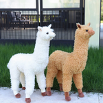 White and brown standing alpaca plushies on grass in front of a black fence