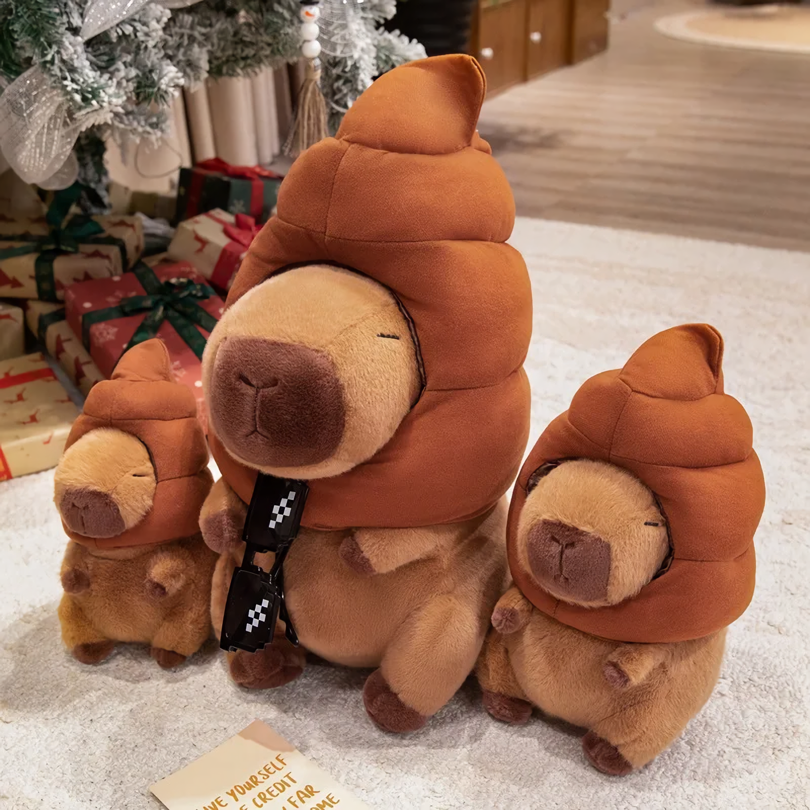 Three capybara plushies with brown swirl hats and sunglasses on a rug beside gifts