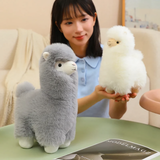 Woman holding a white alpaca plush next to a gray alpaca plush on a table