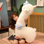 Woman sitting on a bed behind a tall white alpaca plush with a blue bow