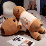 Two lying capybara plush pillows in cream shirts on a floor beside magazines