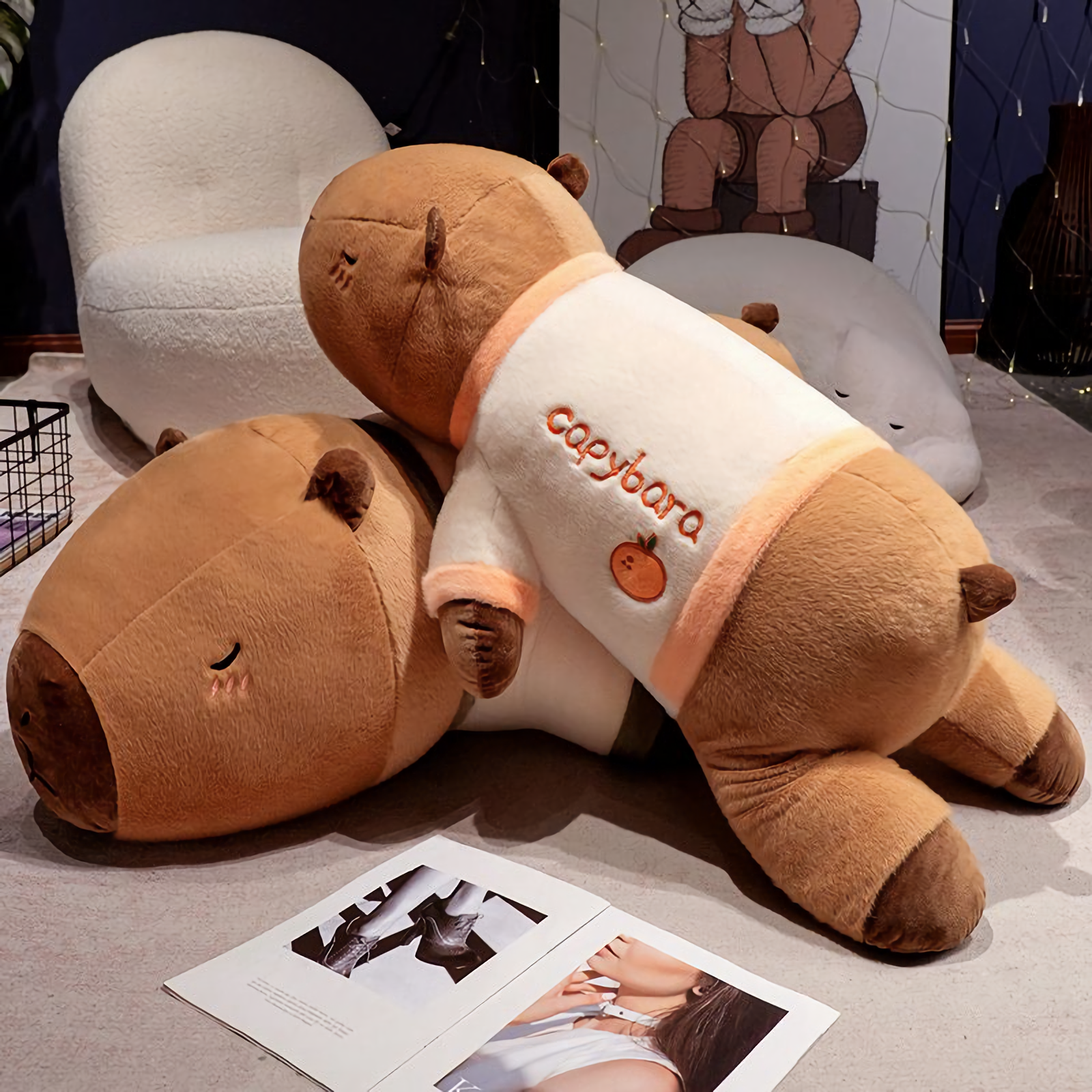 Two lying capybara plush pillows in cream shirts on a floor beside magazines