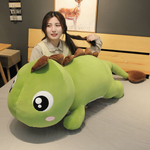 Woman sitting beside a green dinosaur plush pillow with brown spikes on a bed.