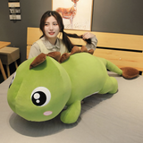 Woman sitting beside a green dinosaur plush pillow with brown spikes on a bed.