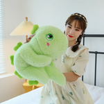 A woman holding a green giant dinosaur plush pillow on a bed.