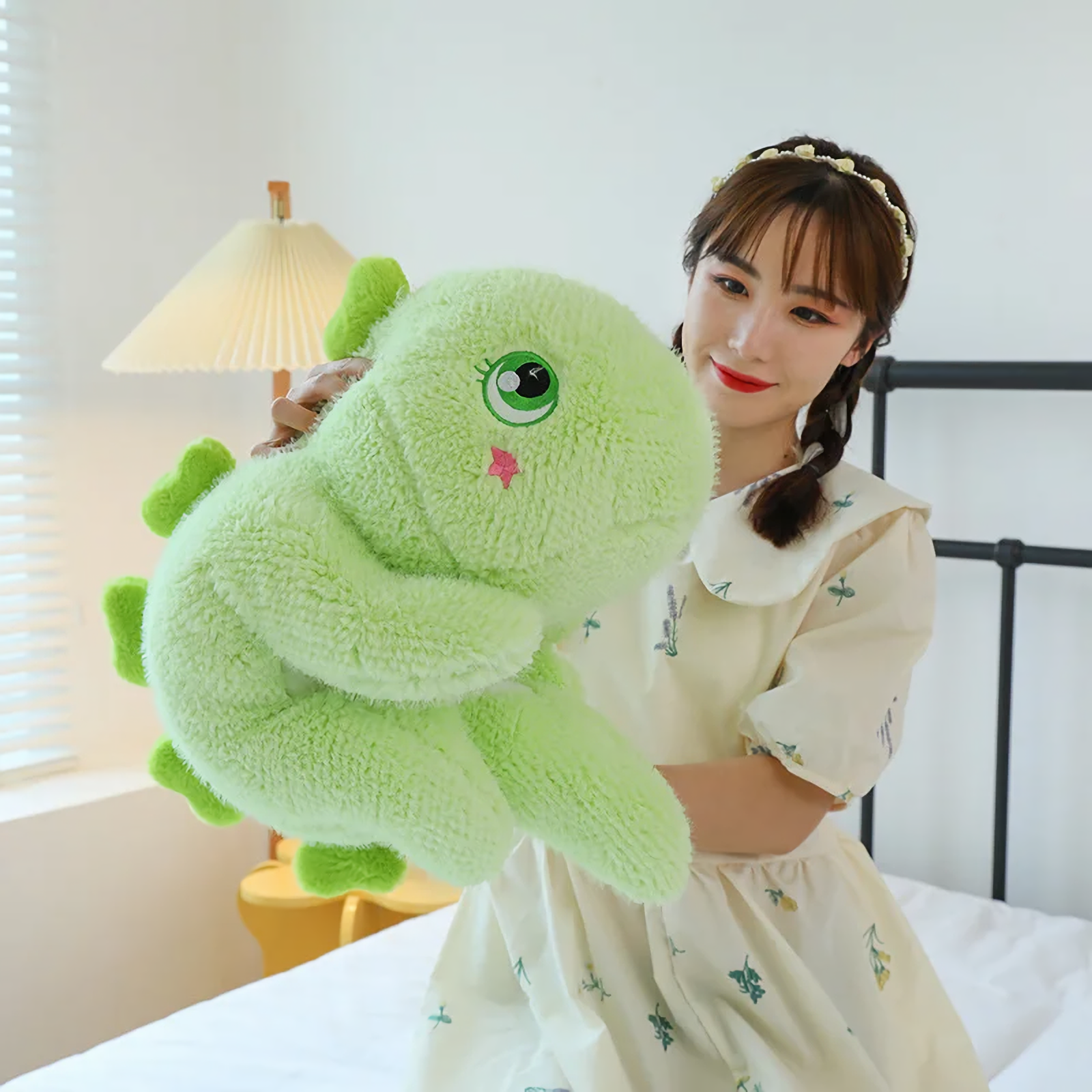 A woman holding a green giant dinosaur plush pillow on a bed.