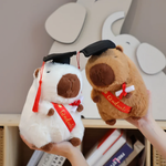 Hands holding a white and a brown graduation capybara plush in front of a shelf