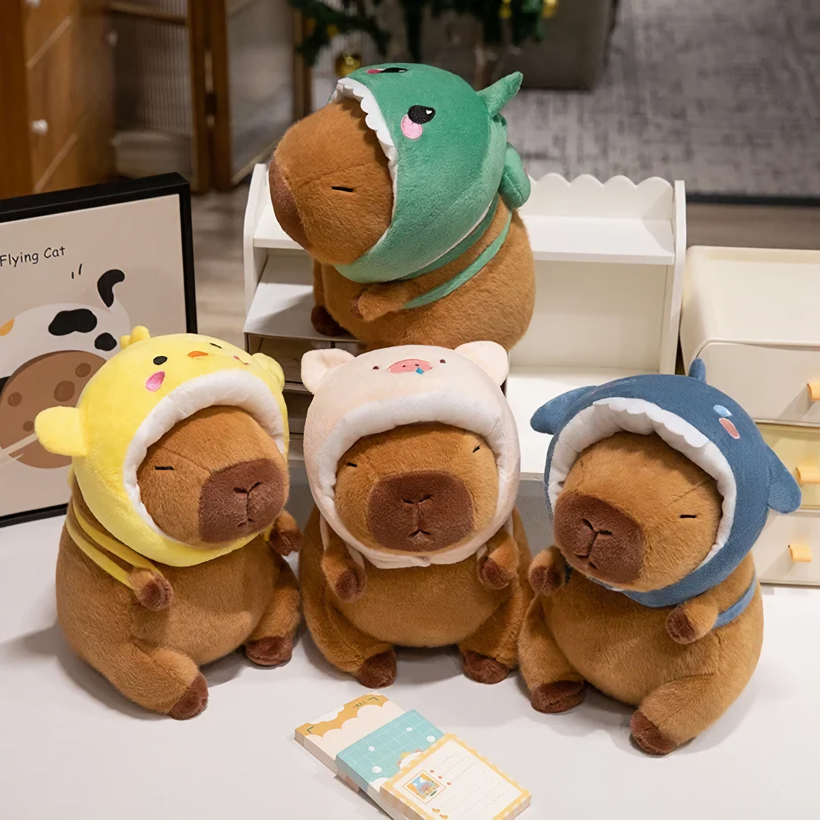 Four capybara plushies in dinosaur, chick, pig and shark hoods on a table