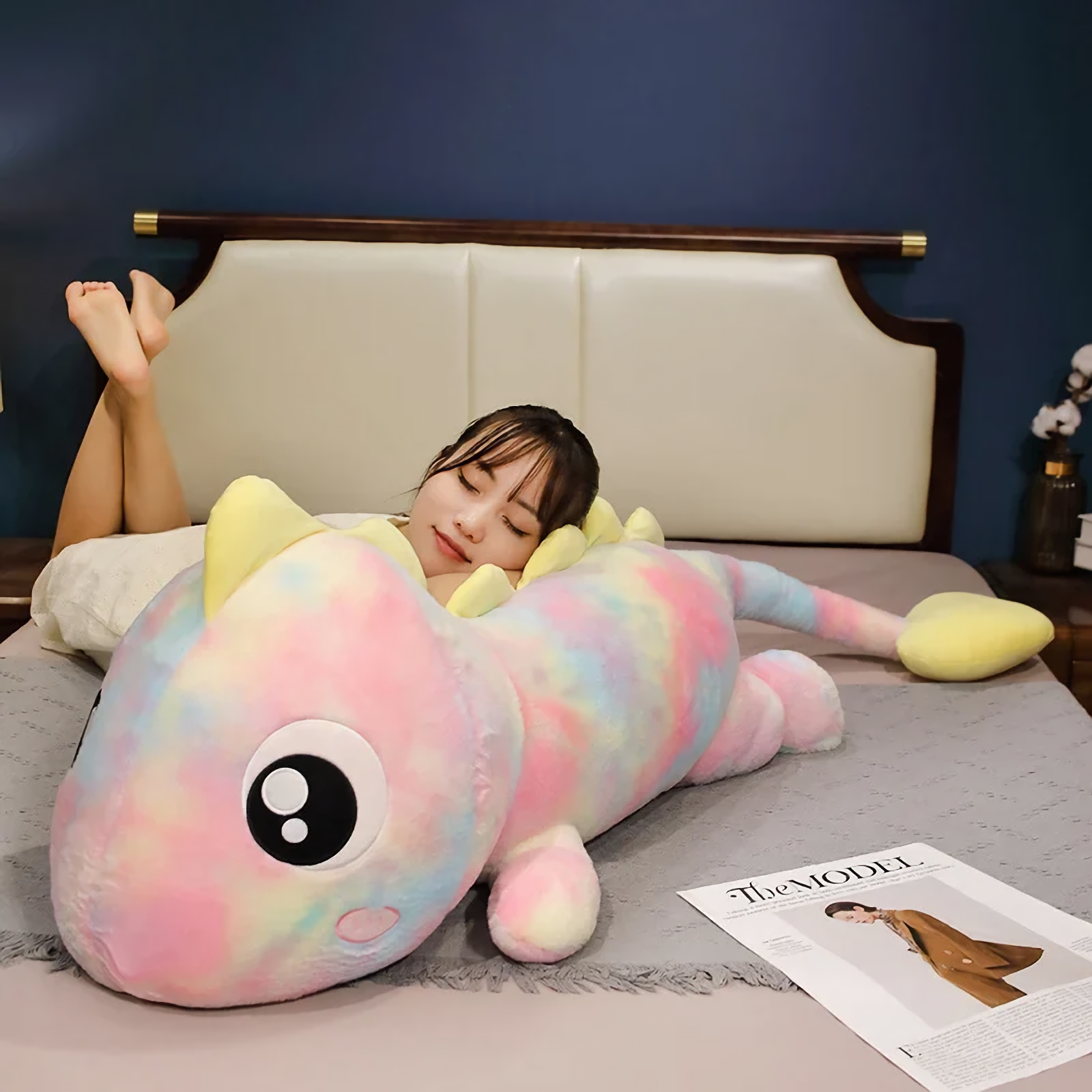Woman lying on a bed resting on a rainbow dinosaur plush body pillow with big eyes