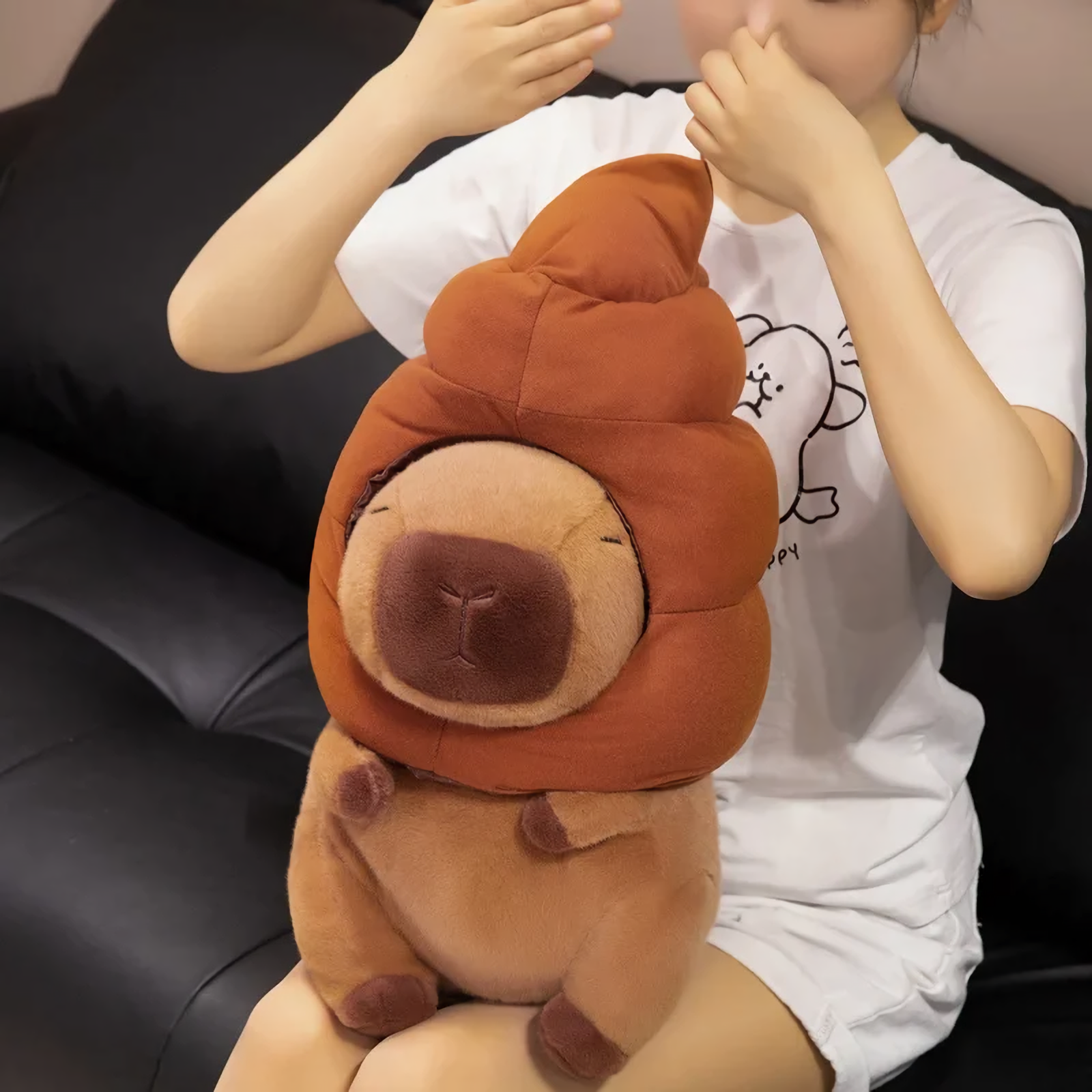 Person sitting on a sofa holding a capybara plush with a brown swirl hat