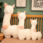 Three white alpaca plushies in different sizes with pink bows and bags on a bed