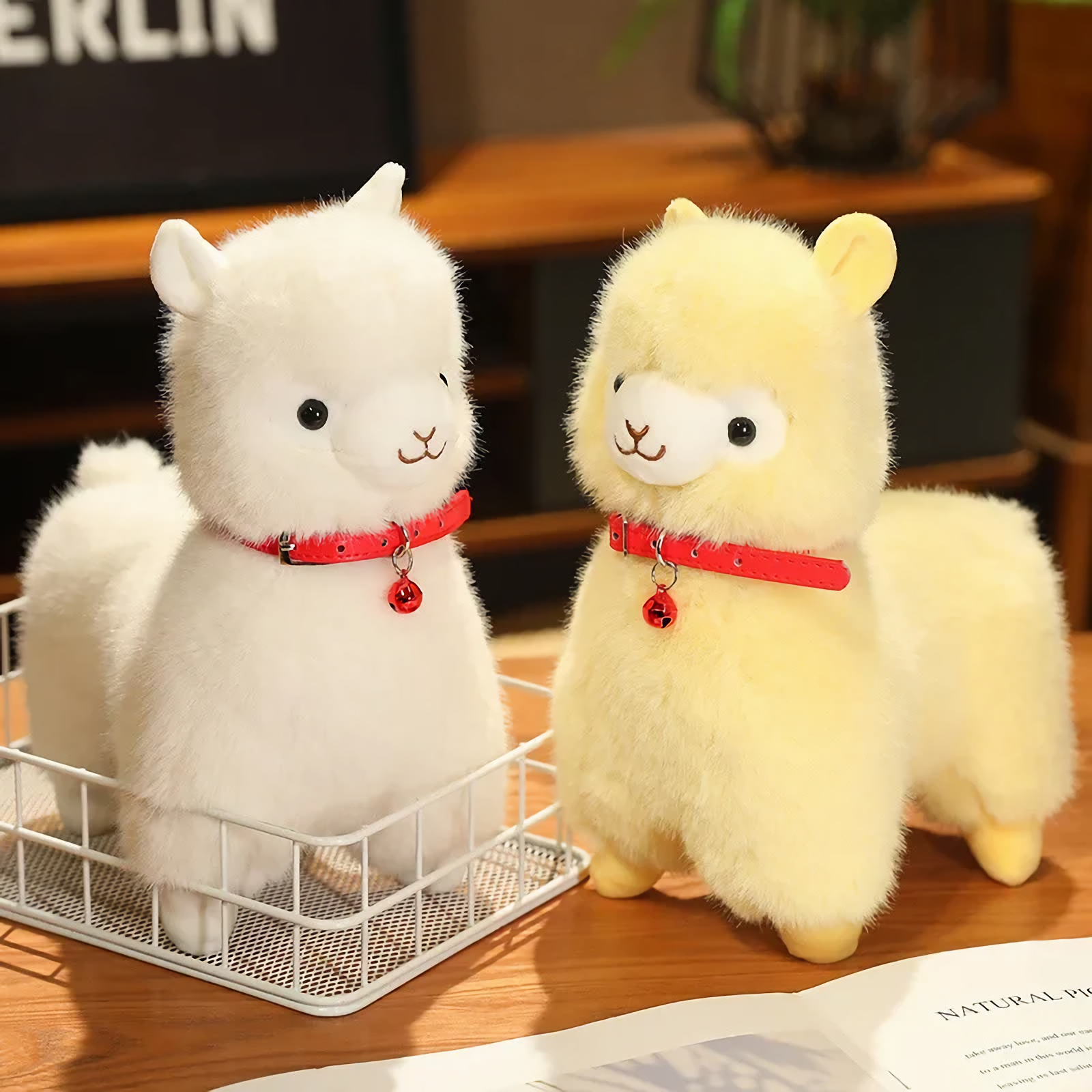 White and yellow alpaca plushies with red bell collars on a table beside a wire basket