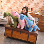 A woman sits on a trunk holding three Brachiosaurus plush toys in green, pink, and blue.
