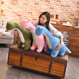A woman sits on a trunk holding three Brachiosaurus plush toys in green, pink, and blue.