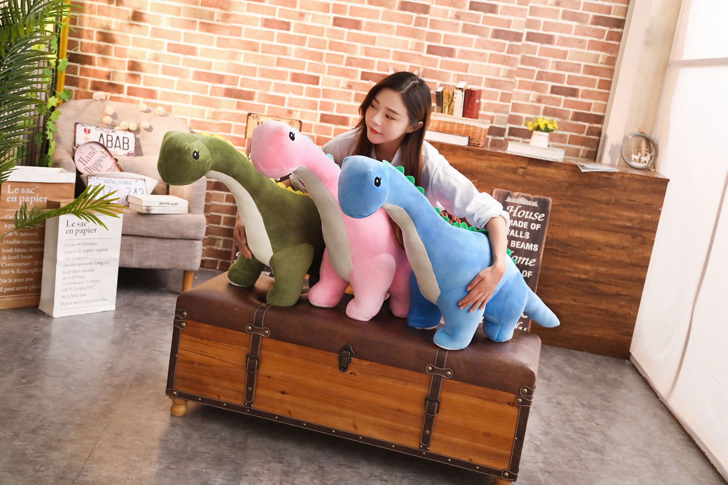 A woman sits on a trunk holding three Brachiosaurus plush toys in green, pink, and blue.