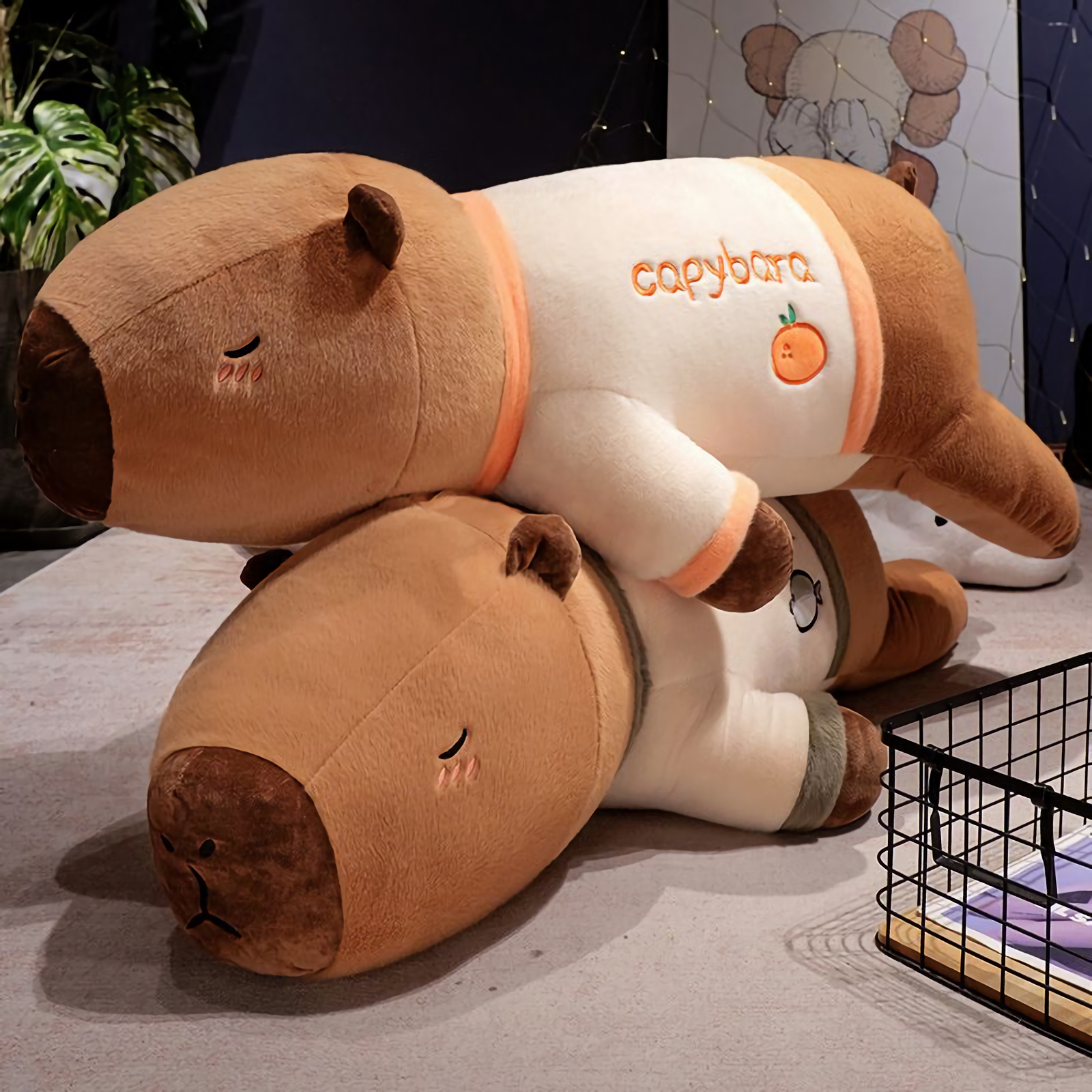 Two lying capybara plush pillows in cream shirts with orange trim on a gray rug