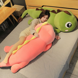 A woman hugging a long pink dinosaur plush pillow on a bed with a green plush pillow beside her.
