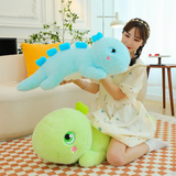 A woman holds a blue giant dinosaur plush pillow, with a green one on the floor nearby.
