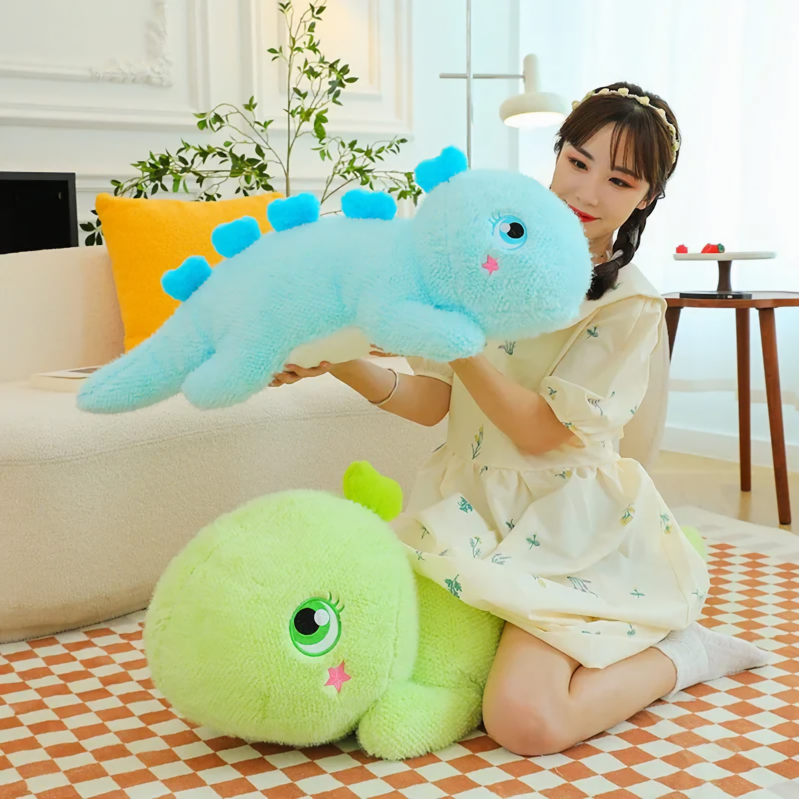 A woman holds a blue giant dinosaur plush pillow, with a green one on the floor nearby.