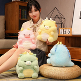 A woman holding a yellow pig-dragon plush, with pink and green pig-dragons beside her on the sofa.