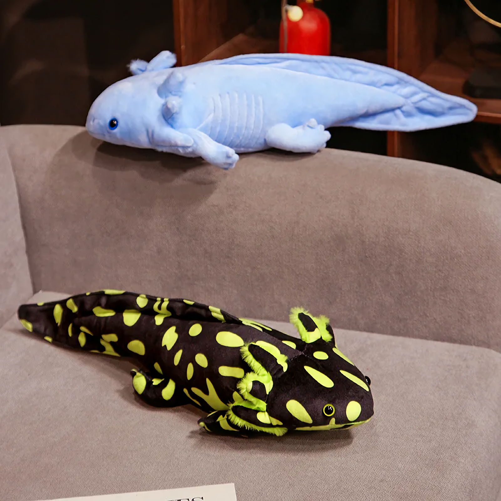 Blue axolotl plush lying on a gray sofa above a black and lime green spotted axolotl plush