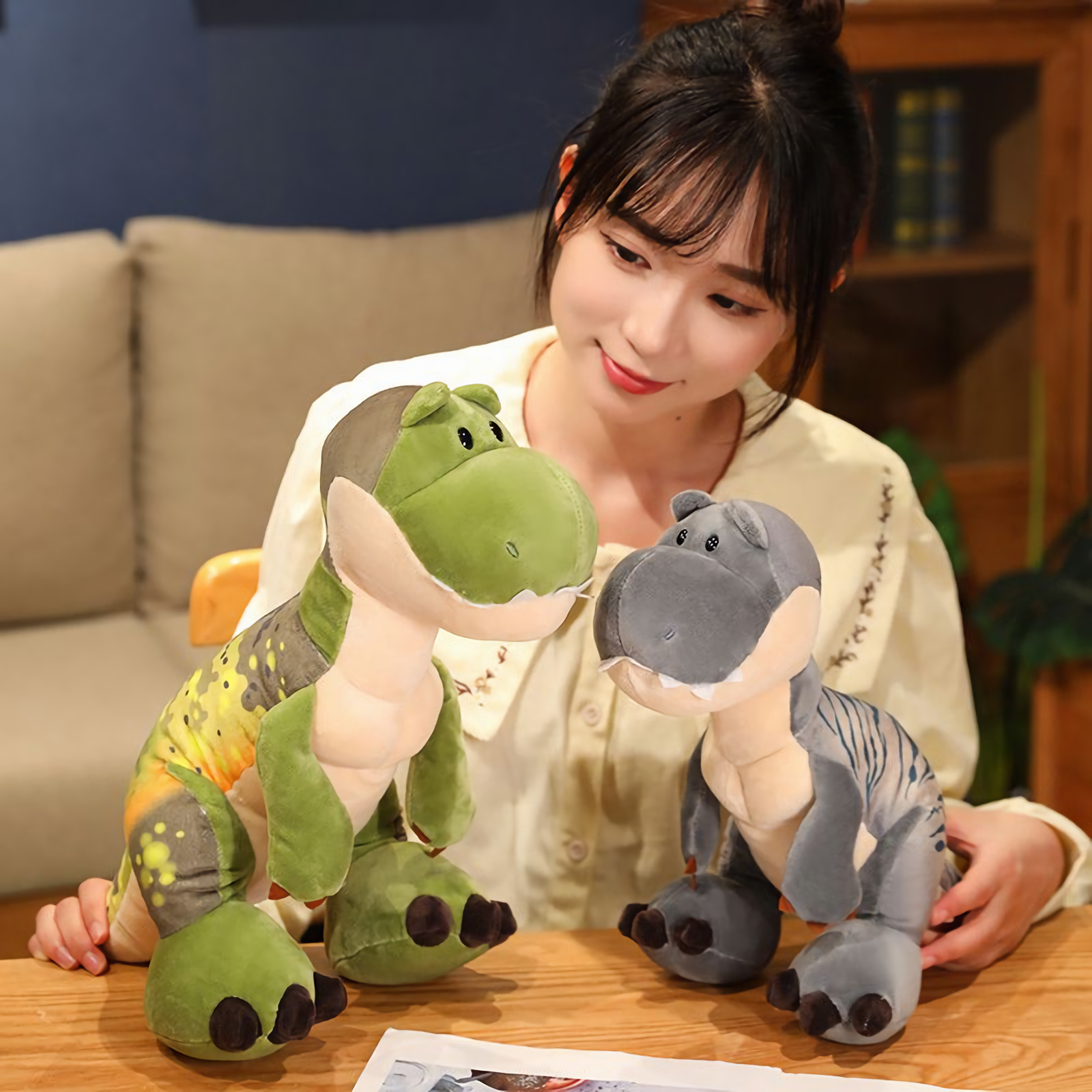 Woman sitting with green and gray T-Rex plush toys on a wooden table, smiling and looking at them.