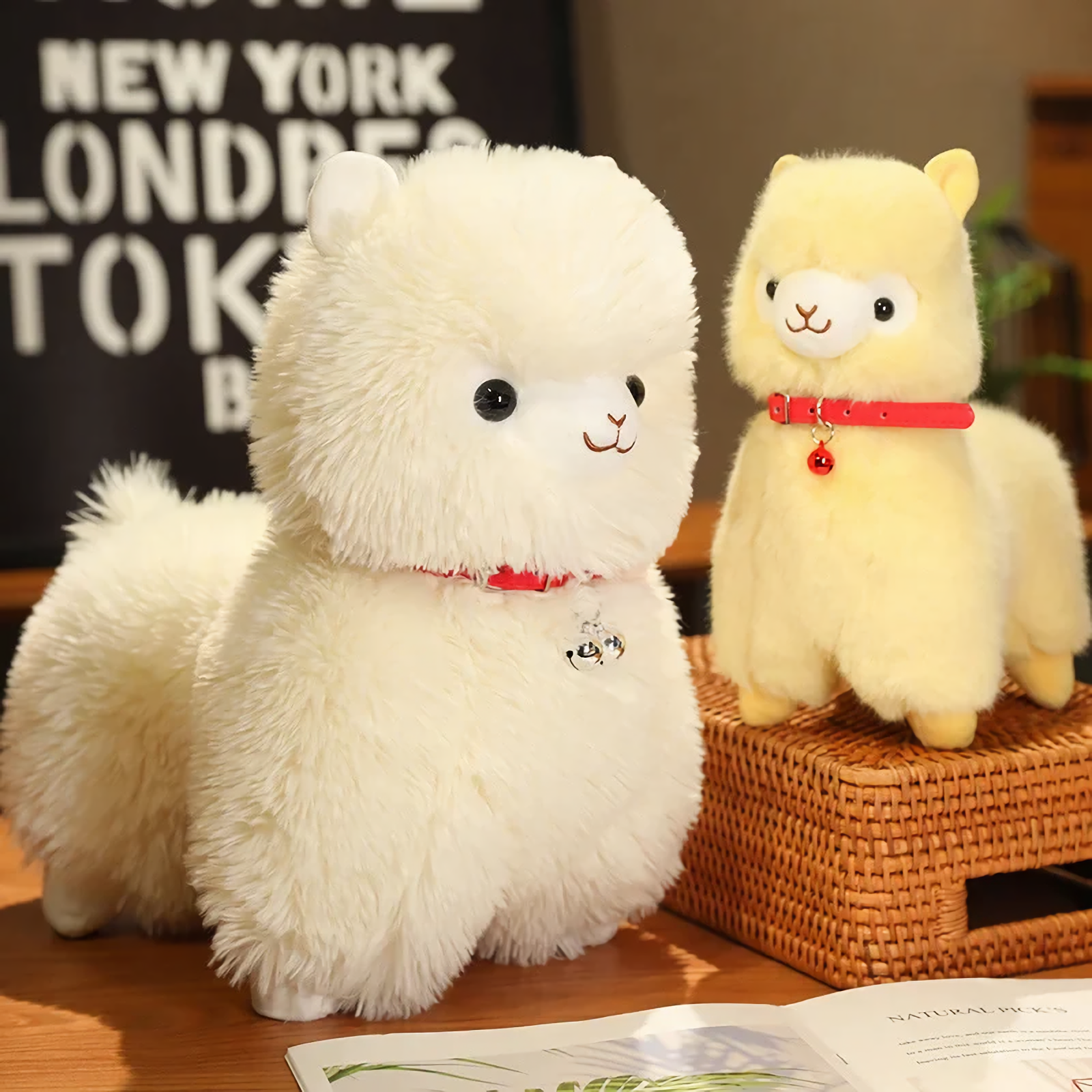 Large white and small yellow alpaca plushies with red bell collars on a wicker stool