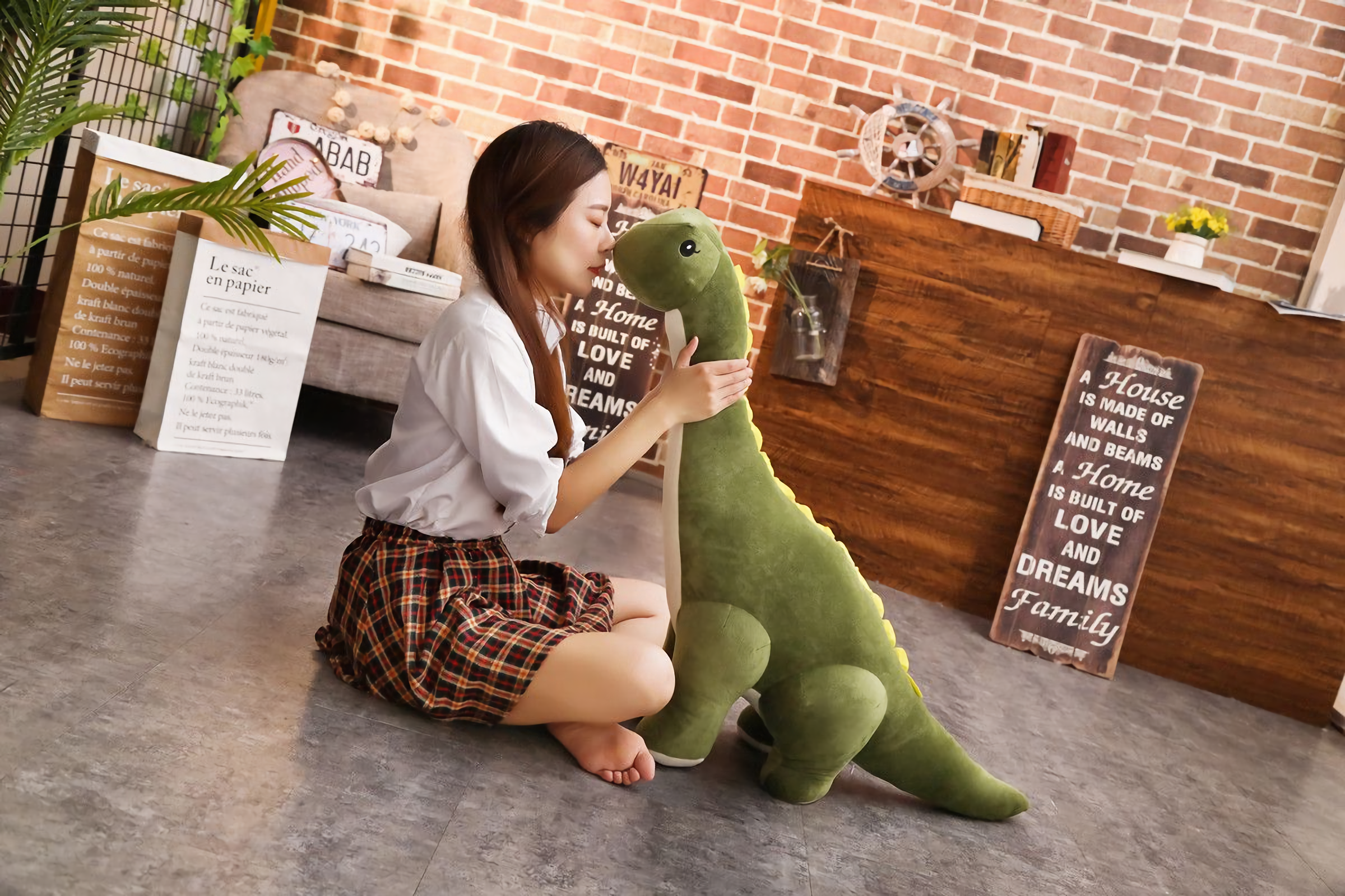 Woman sitting on the floor hugging a green Brachiosaurus dinosaur plush with cream belly and back spikes.