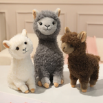 White, gray and brown fluffy alpaca plushies sitting together on a bed