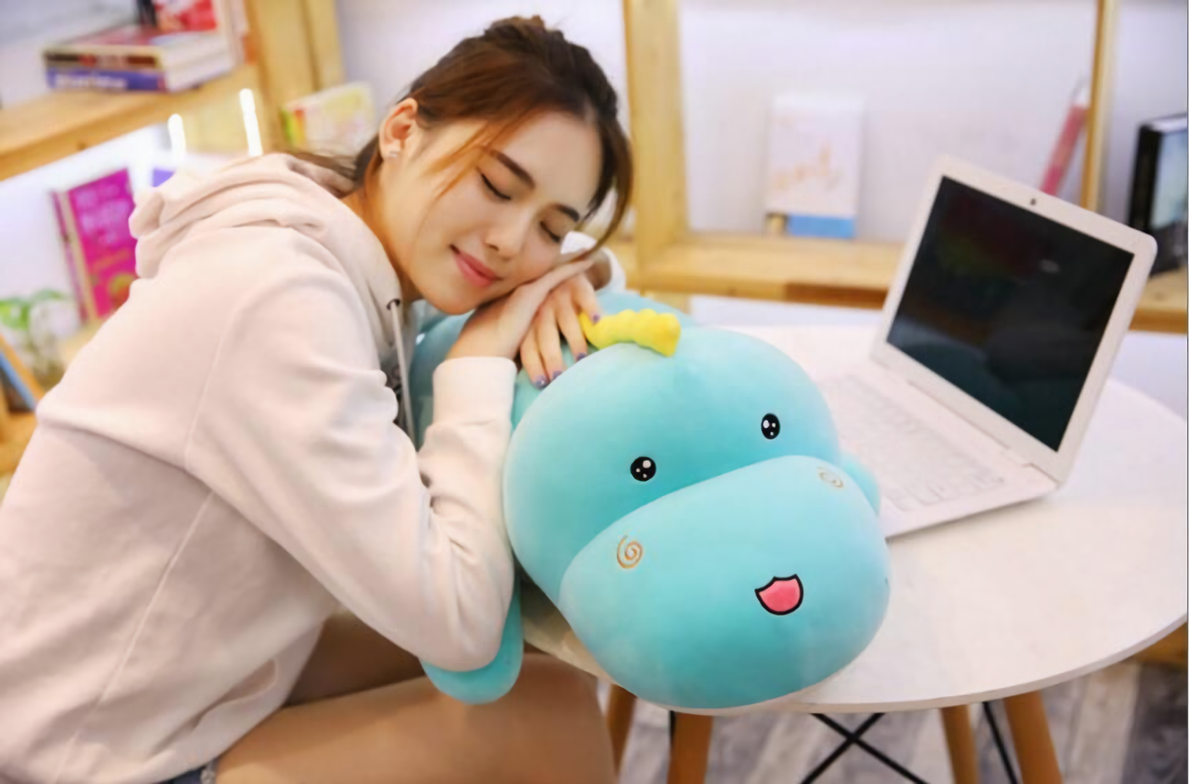 Woman resting her head on a blue dinosaur plush pillow beside an open laptop
