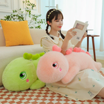 A woman reads on a sofa with pink giant dinosaur plush on lap and green plush beside her.