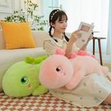 A woman reads on a sofa with pink giant dinosaur plush on lap and green plush beside her.